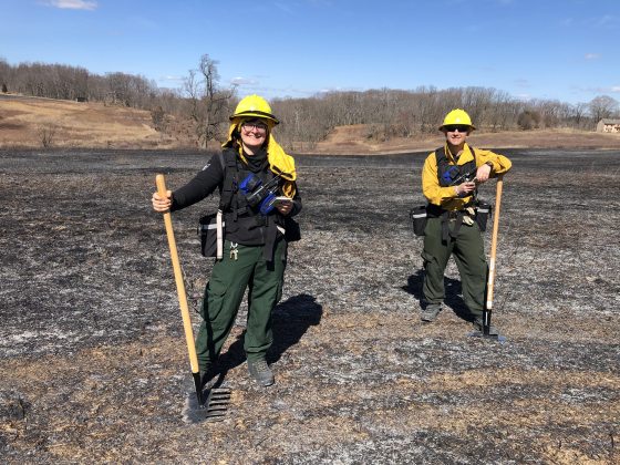 Two people standing in a burned meadow in protective gear.